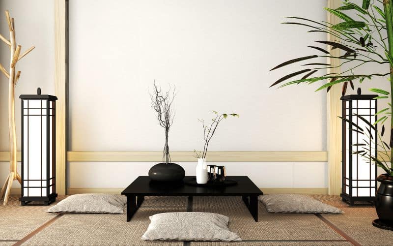 Salon asiatique, avec une table basse noire avec quatre coussins de sol disposés autour, une lanterne en bois et noir et papier de chaque côté, une plante verte et un mur blanc dans le fond.