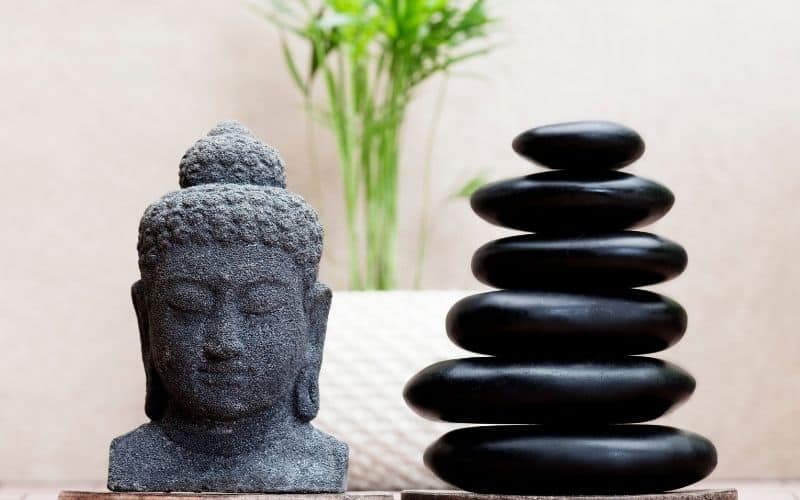 Tête de Bouddha en pierre, posée sur un meuble avec des galets noir empilés à côté et un pot blanc avec une plante verte derrière.